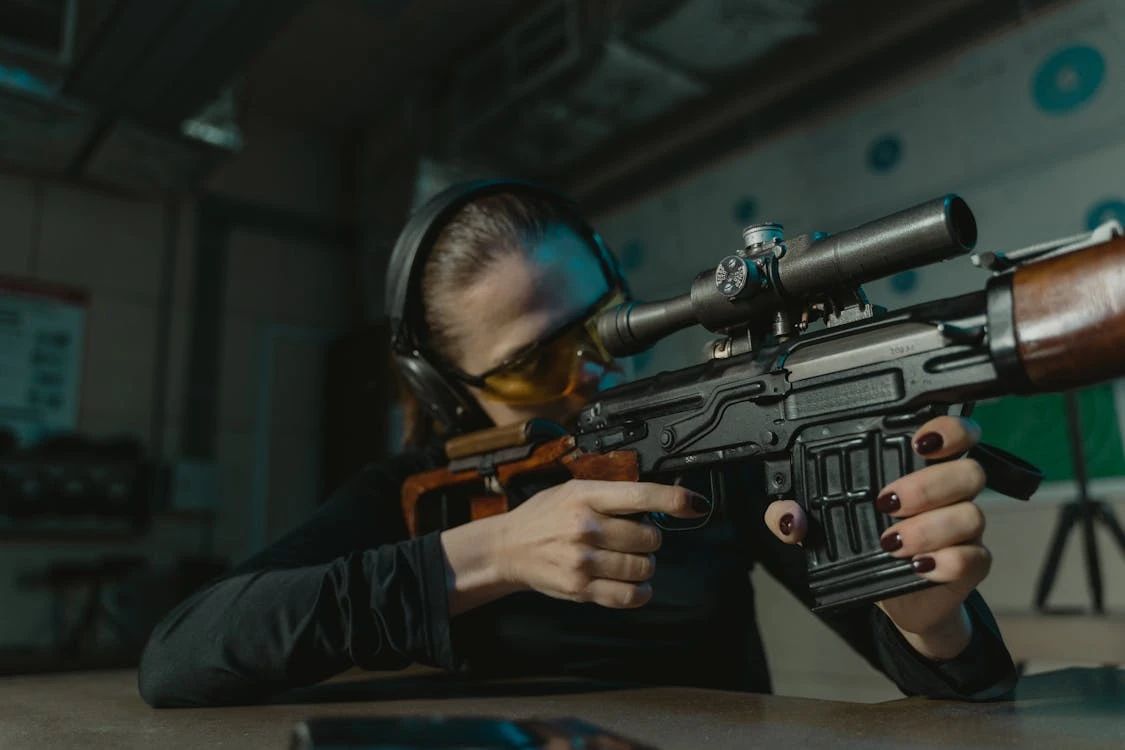 Free Woman in shooting range, wearing ear and eye protection, aiming a rifle with focus. Stock Photo